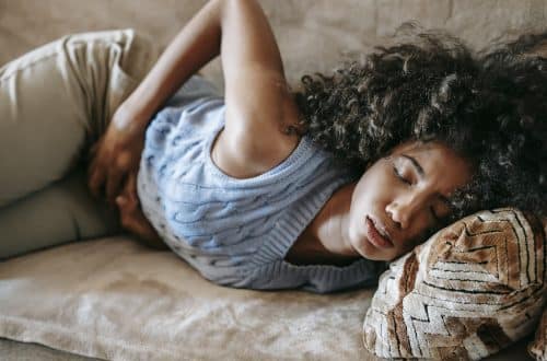 High angle of African American woman with afro hairstyle and closed eyes having pain in belly lying on sofa